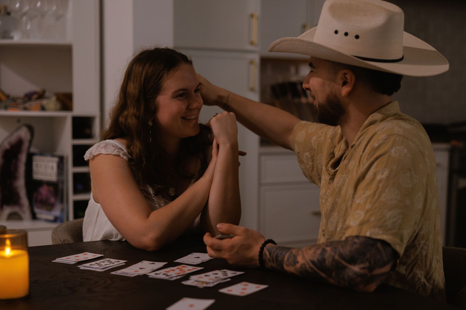 Tattooed man in cowboy hat playfully touching woman's forehead by candlelight with tarot cards.