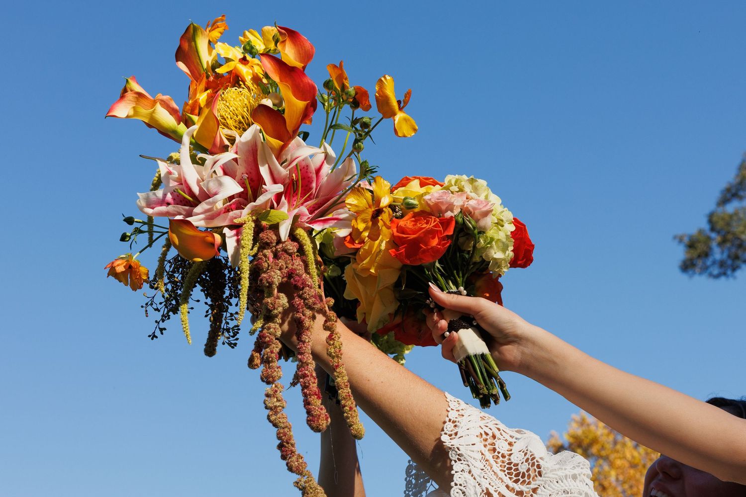 Bride and bridesmaid holding colorful fall wedding bouquets with lilies and roses against a clear blue sky.