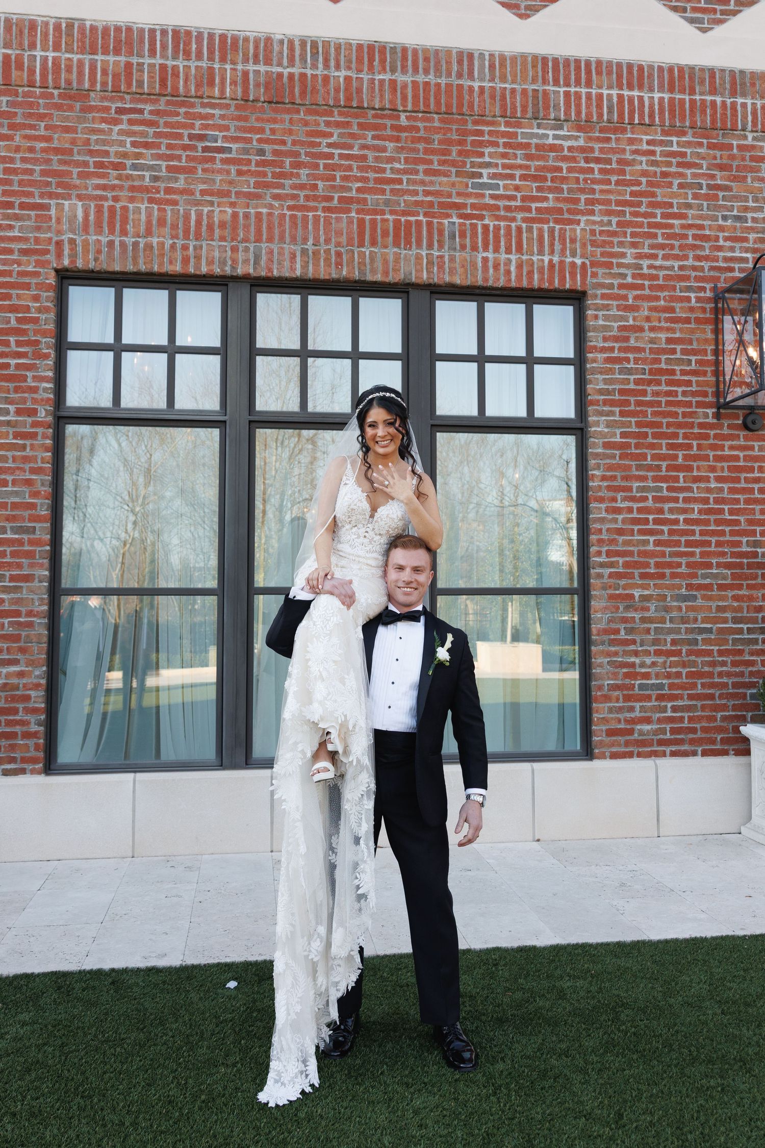 Groom lifts bride onto his shoulders in front of a brick building exterior at their wedding venue at The Yorkmont in Charlotte, NC