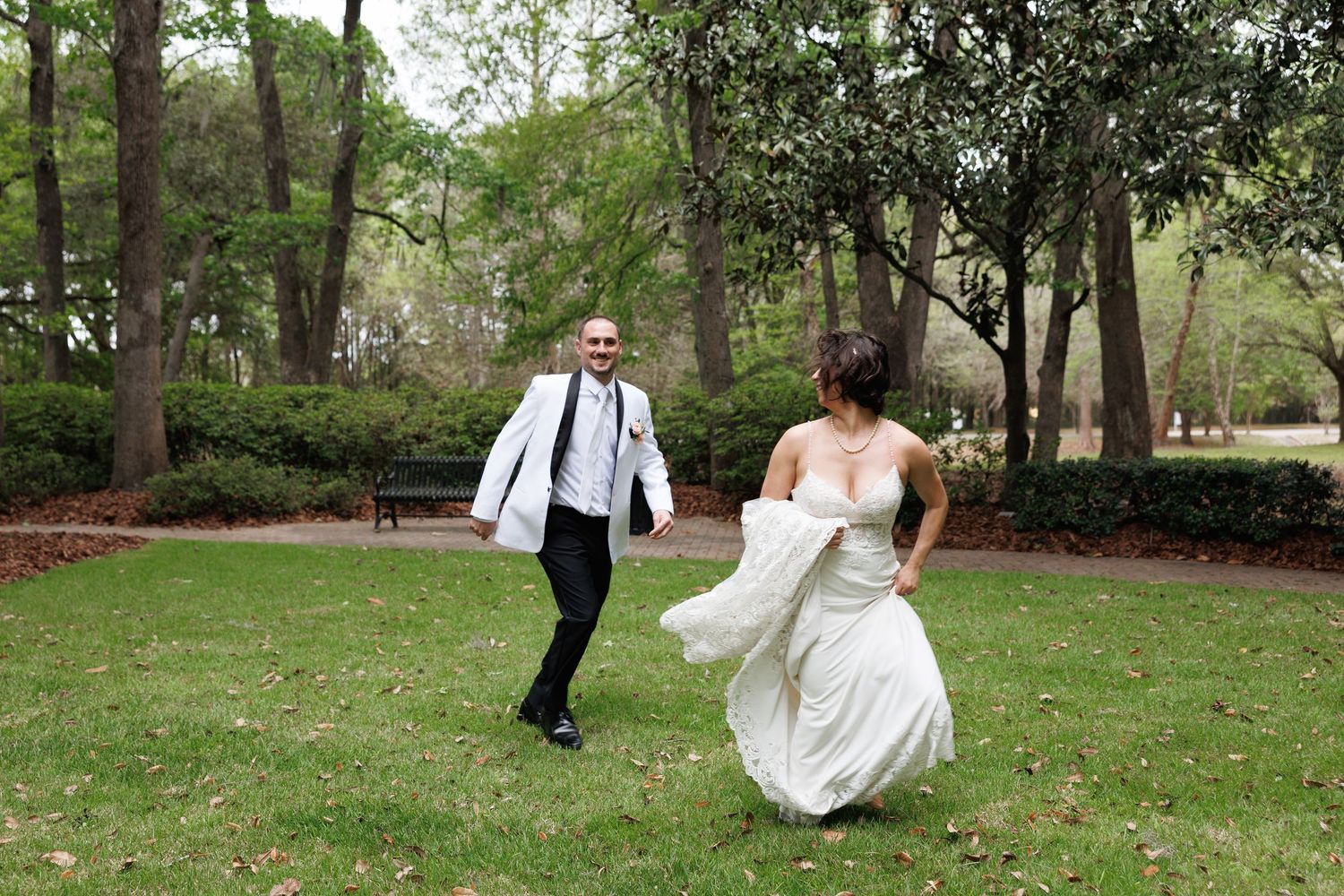 Bride and groom playfully run across green lawn in park during outdoor wedding photo session.