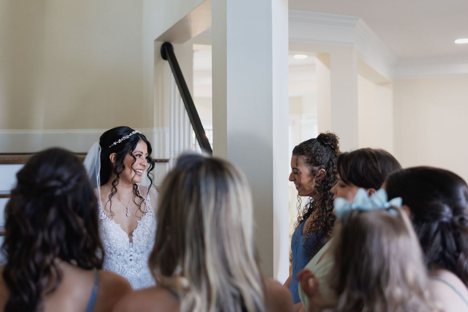 Bride in white lace gown smiles at bridesmaids on staircase during elegant wedding preparation moment.