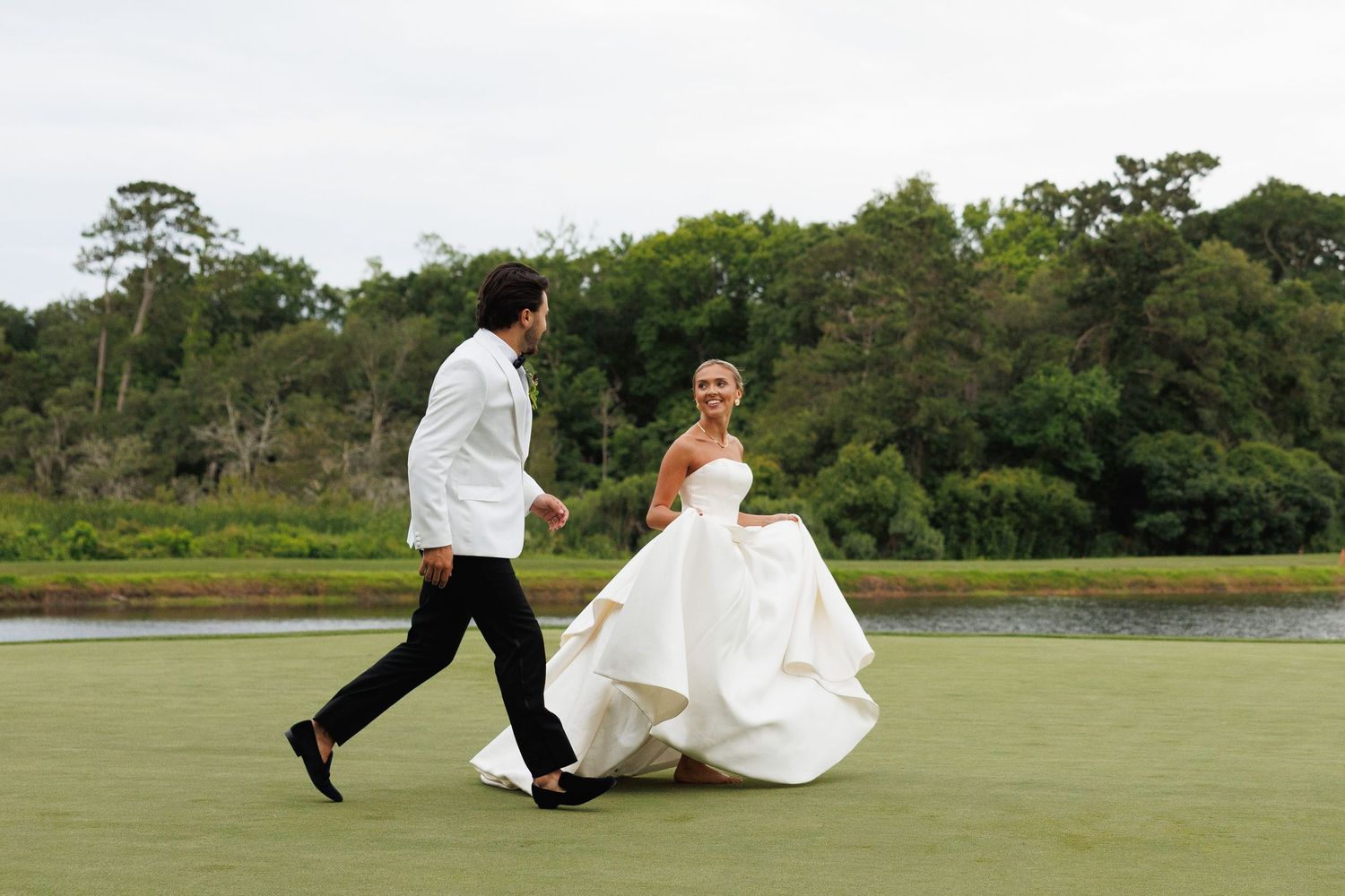Bride in white gown and groom in white jacket walk together on green lawn with trees and water in background.