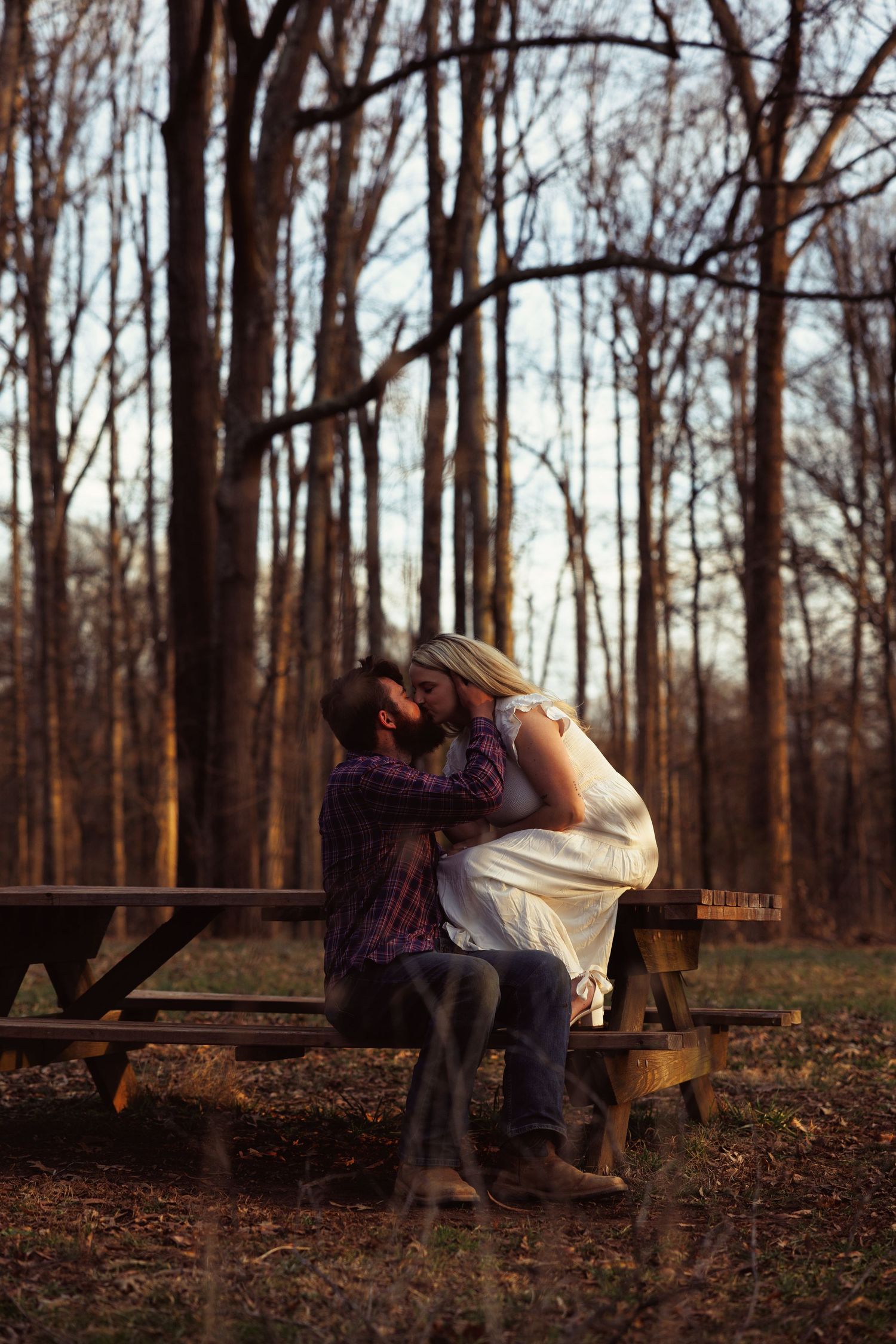 Couple sharing a romantic kiss on a wooden picnic table in a serene forest setting during golden hour.