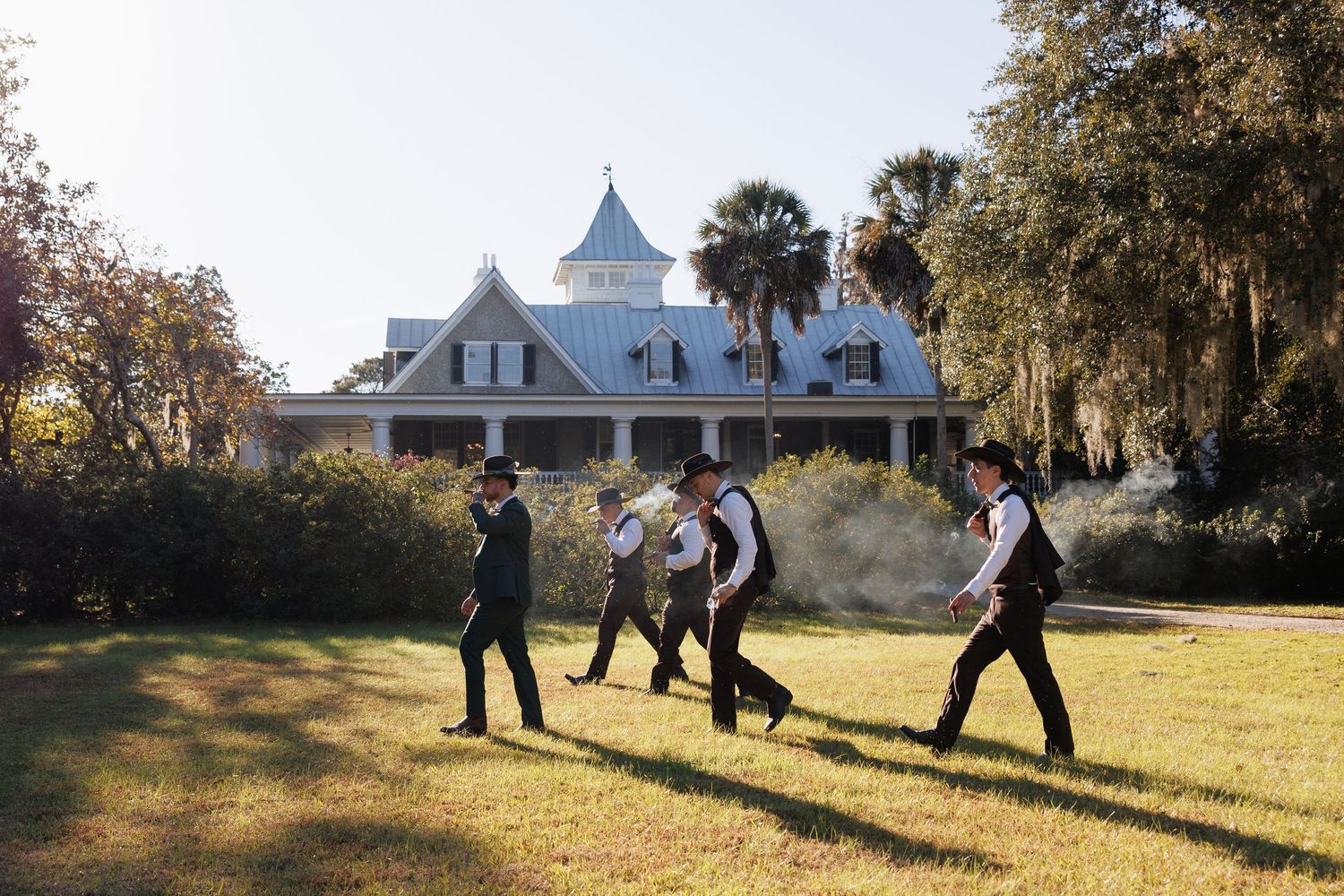 Groomsmen in dark suits walk across lawn in front of historic Southern plantation wedding venue.