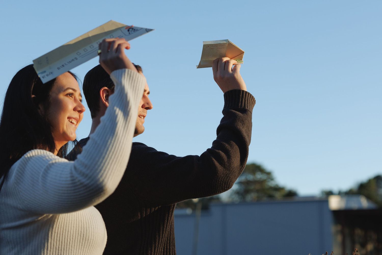 A man and woman hold paper airplanes up against a clear blue sky outdoors.