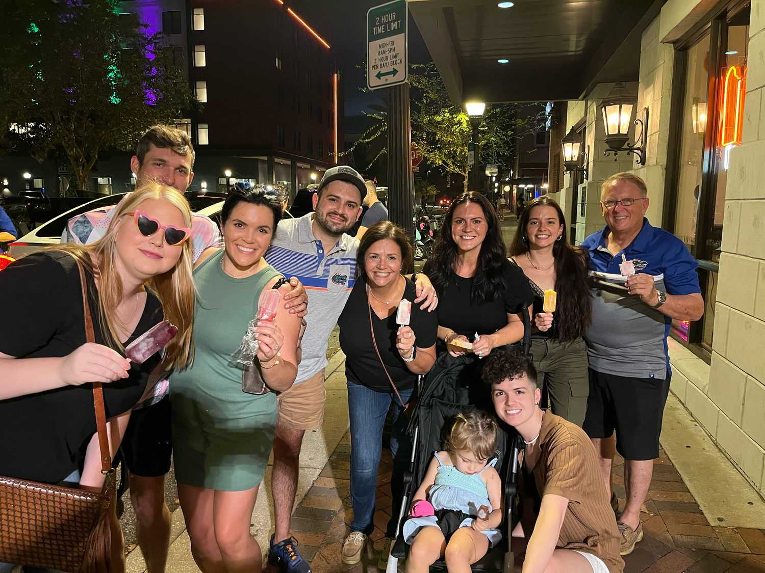 A group of friends and family smiling together on a city sidewalk at night, some holding treats near a lit storefront.