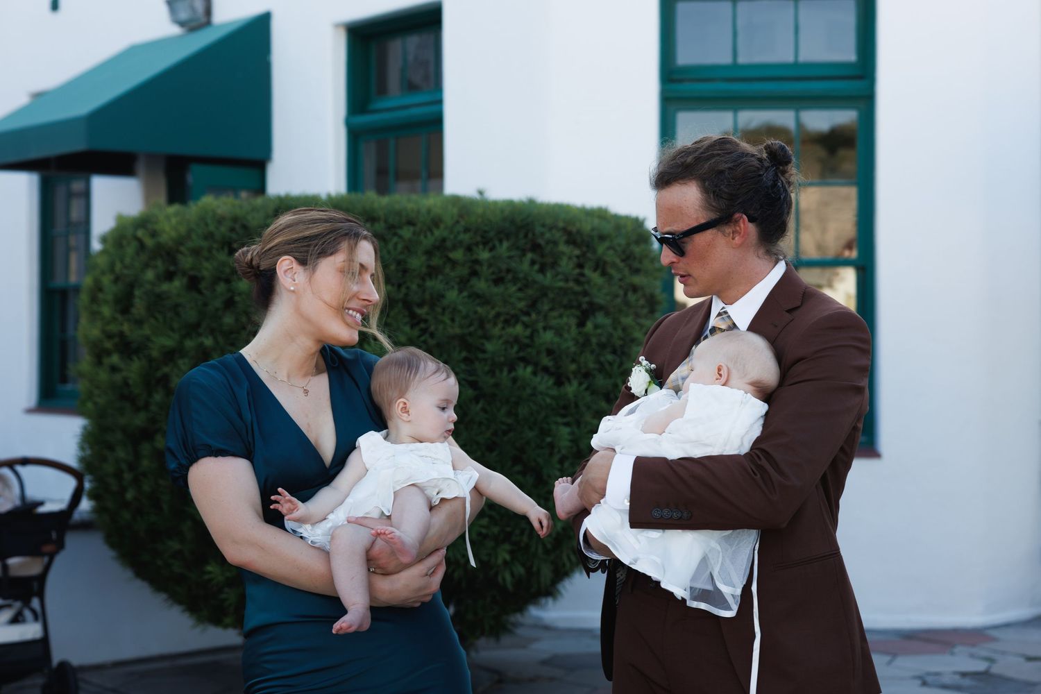Two guests holding babies dressed in white at an outdoor wedding reception near a white building.
