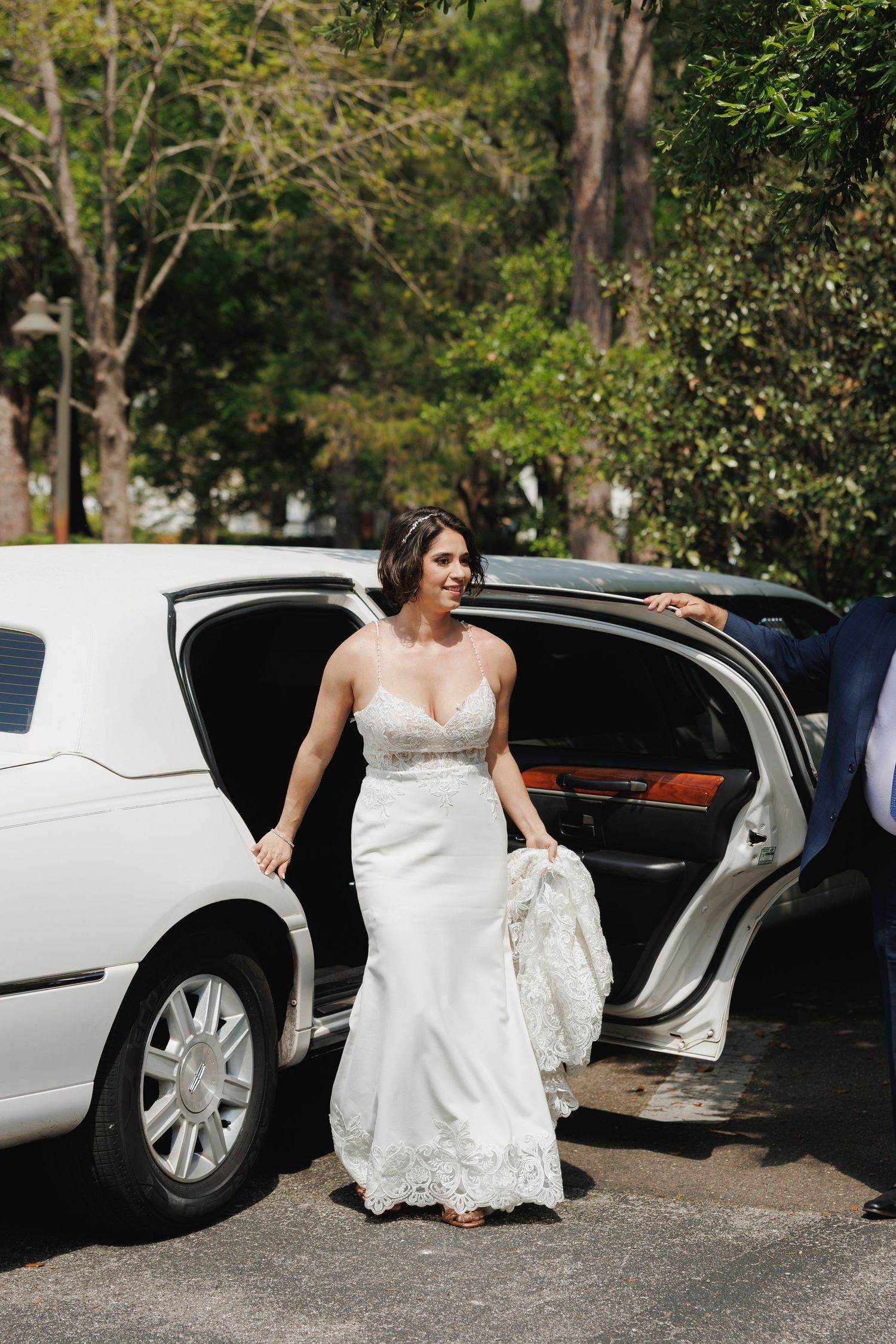 Bride in lace wedding gown stepping out of white limousine surrounded by green trees.