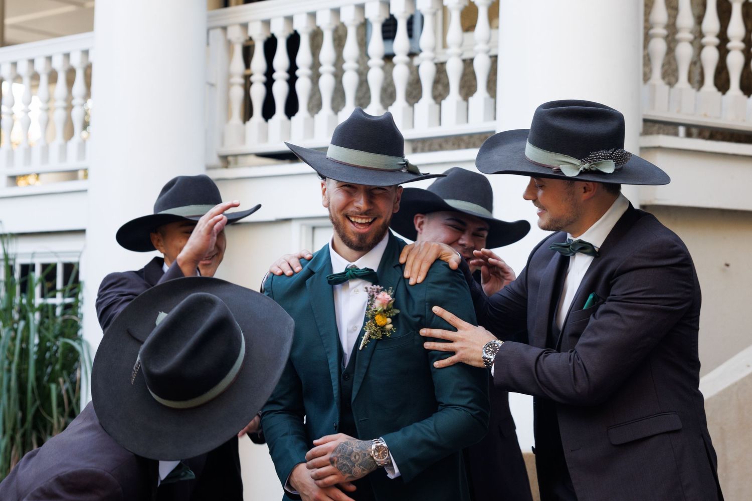 Groom in green suit surrounded by groomsmen in dark suits and wide-brim hats, laughing together outdoors.