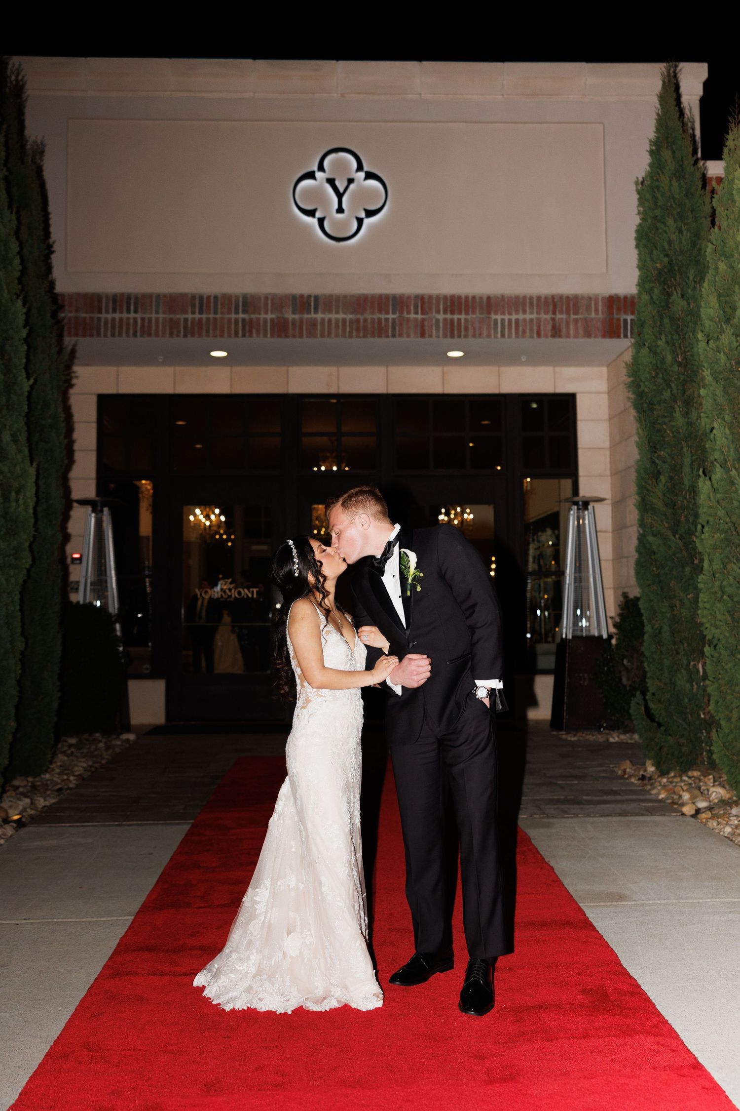 Bride and groom kiss on red carpet beneath glowing venue sign at The Yorkmont in Charlotte, NC flanked by tall cypress trees at night.