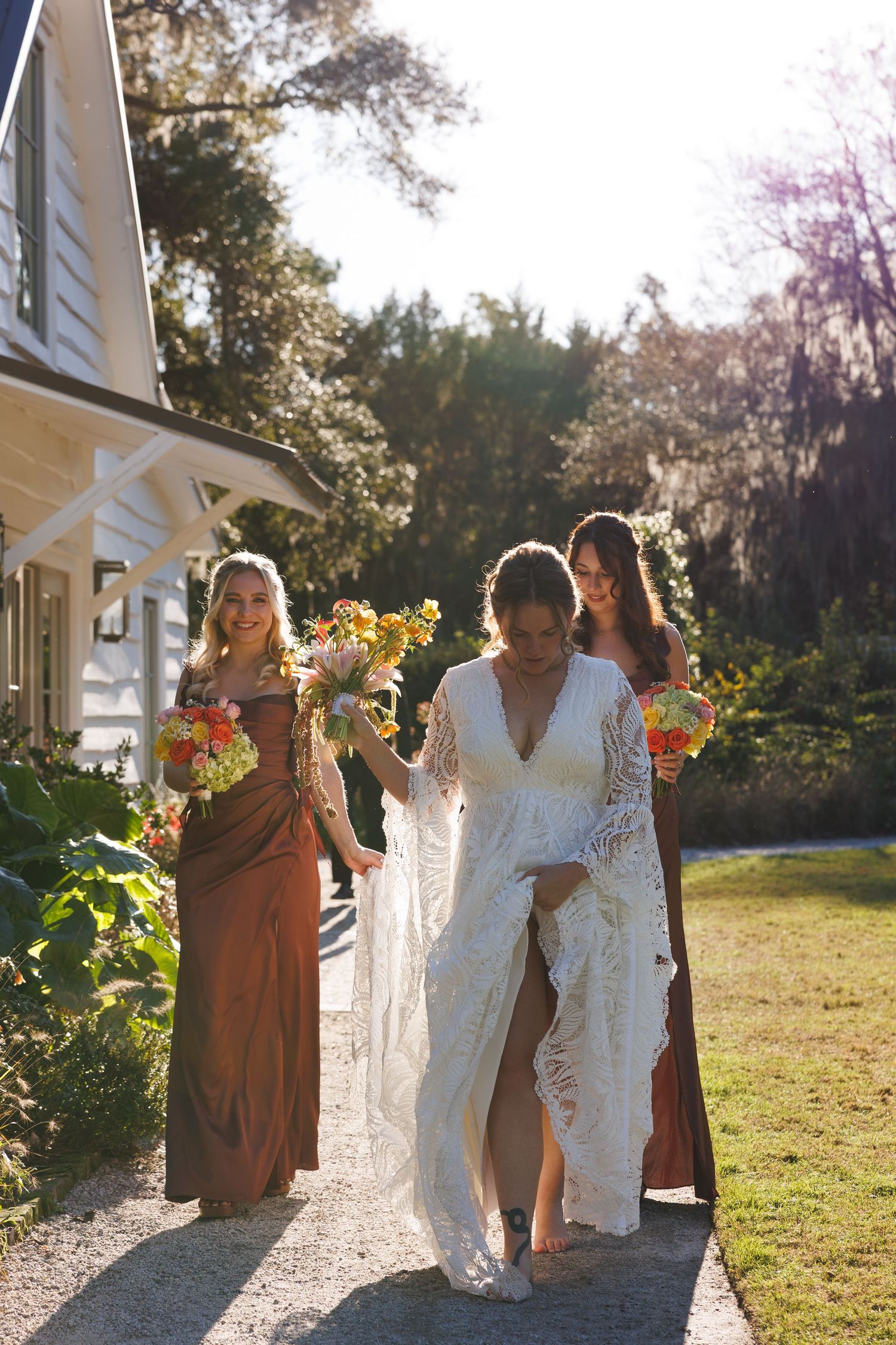 Bride in lace gown walks with two bridesmaids in brown dresses holding colorful bouquets outdoors.