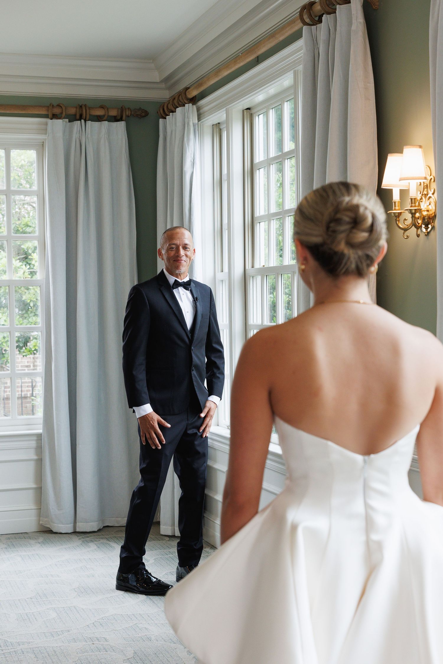 Groom in black tuxedo sees bride in strapless white gown during emotional first look in elegant room.