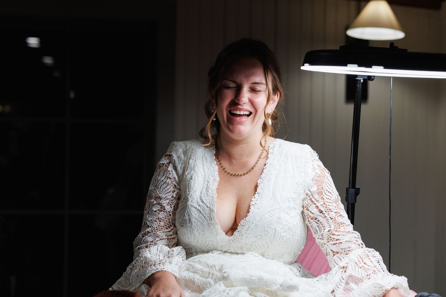 Laughing bride in white lace wedding dress sitting in bright light during wedding preparations.