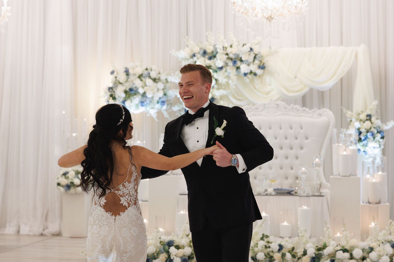 Bride and groom share first dance at elegant wedding reception with white floral decor and draped backdrop.
