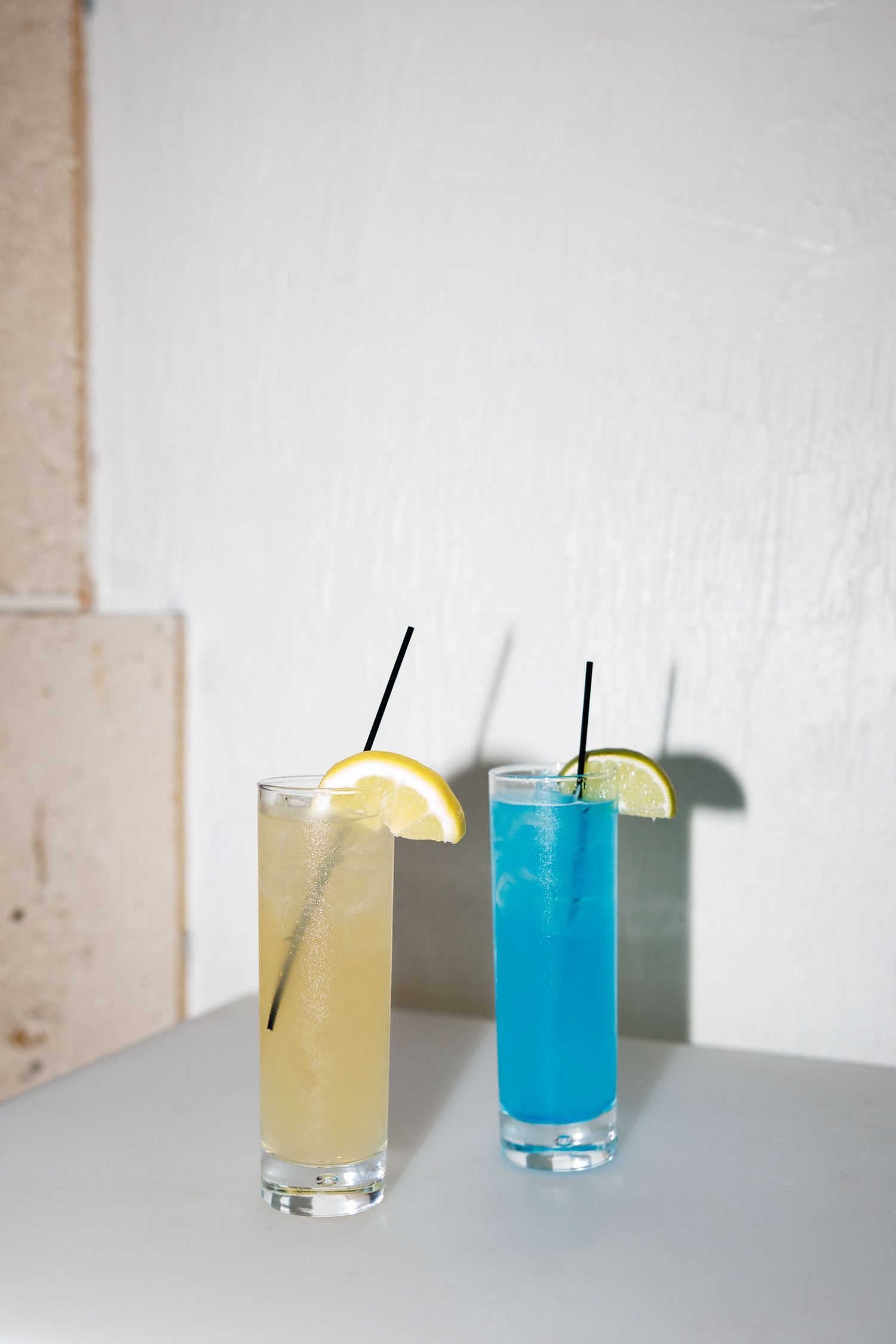 Two tall cocktail glasses with black straws, one yellow and one blue, garnished with citrus slices on a white surface.