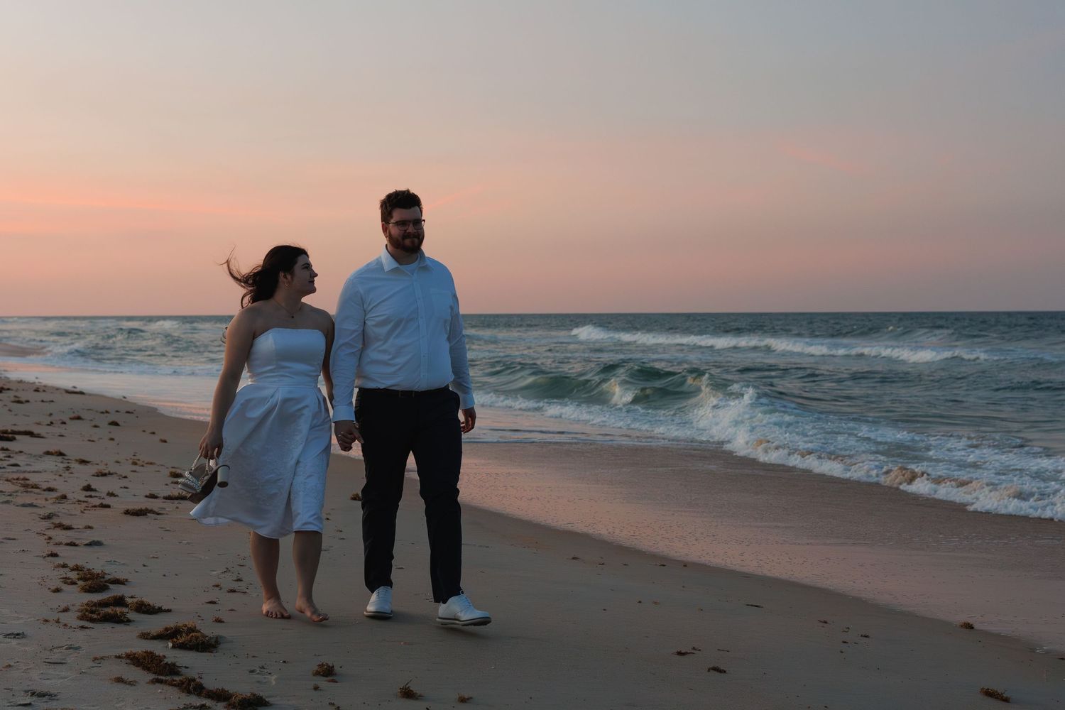 Couple holding hands walking along a sandy beach at sunset with gentle ocean waves nearby.