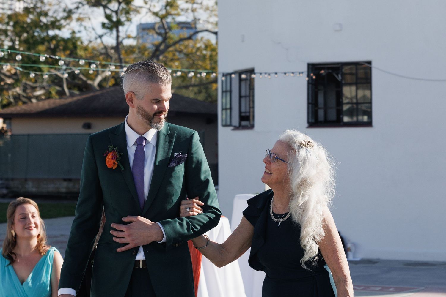 A groom in a green suit walks with an older woman outdoors at a sunlit wedding venue.