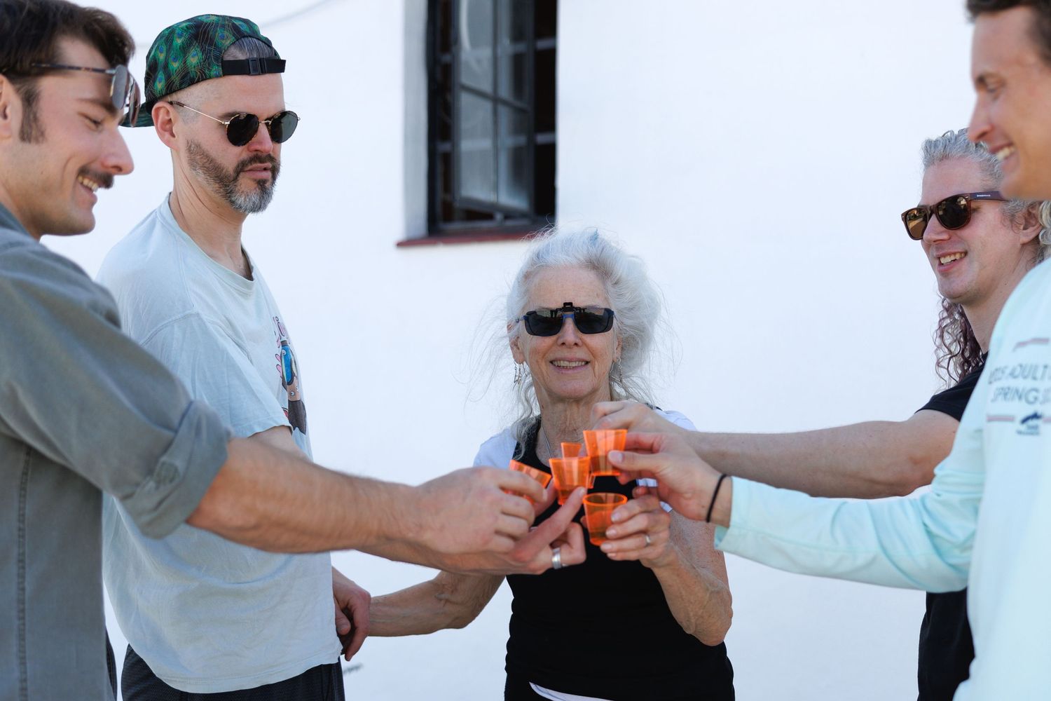 Group of five people toasting with orange drinks outdoors, celebrating together against a white building.