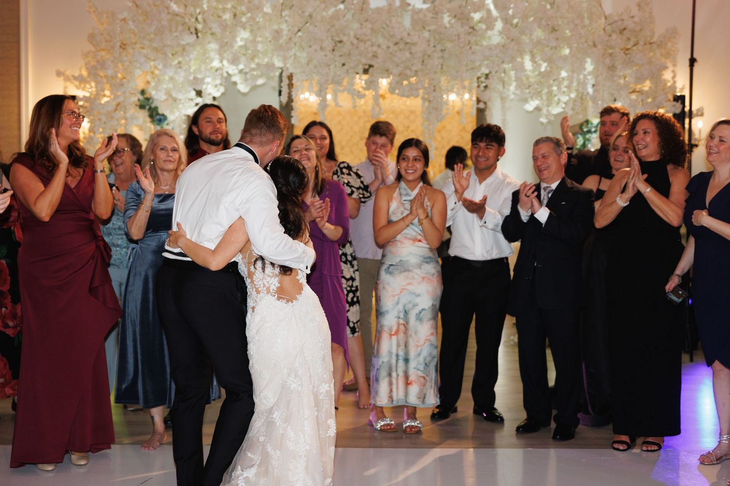 Bride and groom share first dance surrounded by clapping wedding guests at elegant floral-decorated reception venue.