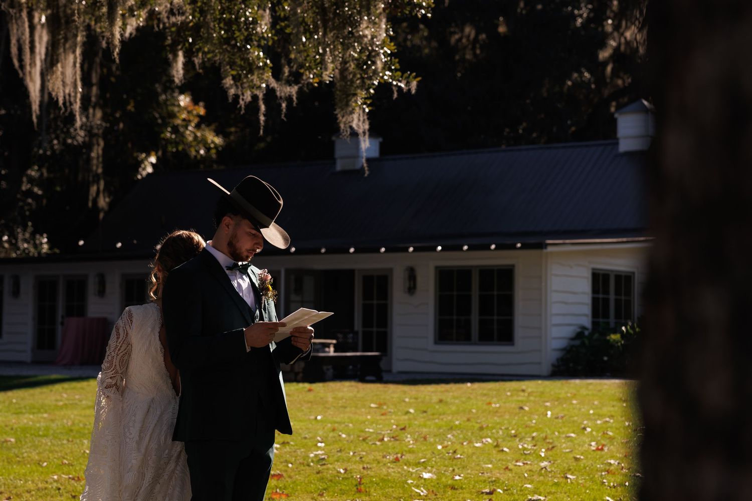Bride and groom share vows outdoors on sunny lawn with Spanish moss trees and white farmhouse backdrop.