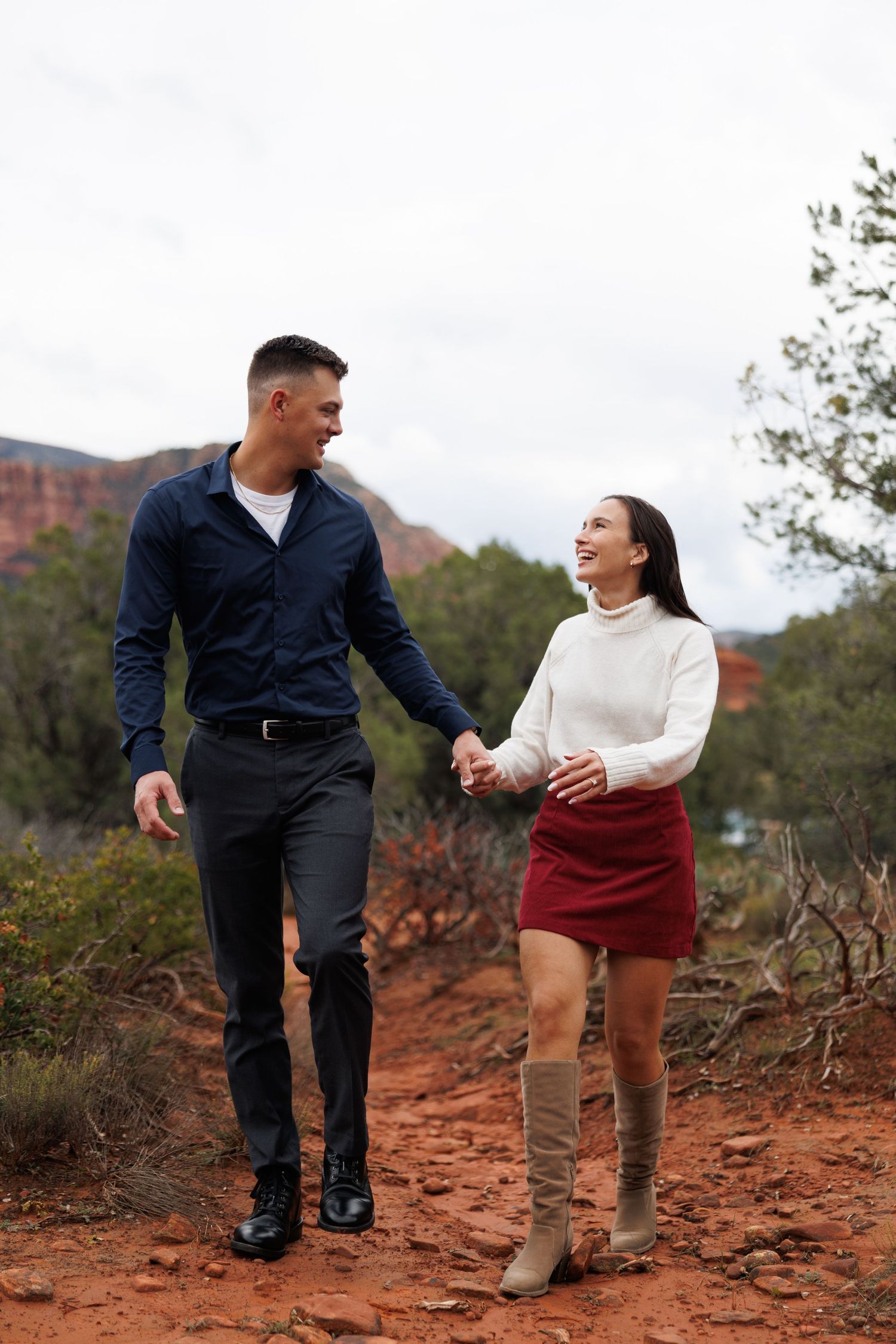Couple holding hands walking on red rock trail in Sedona, Arizona, smiling at each other in stylish fall outfits.