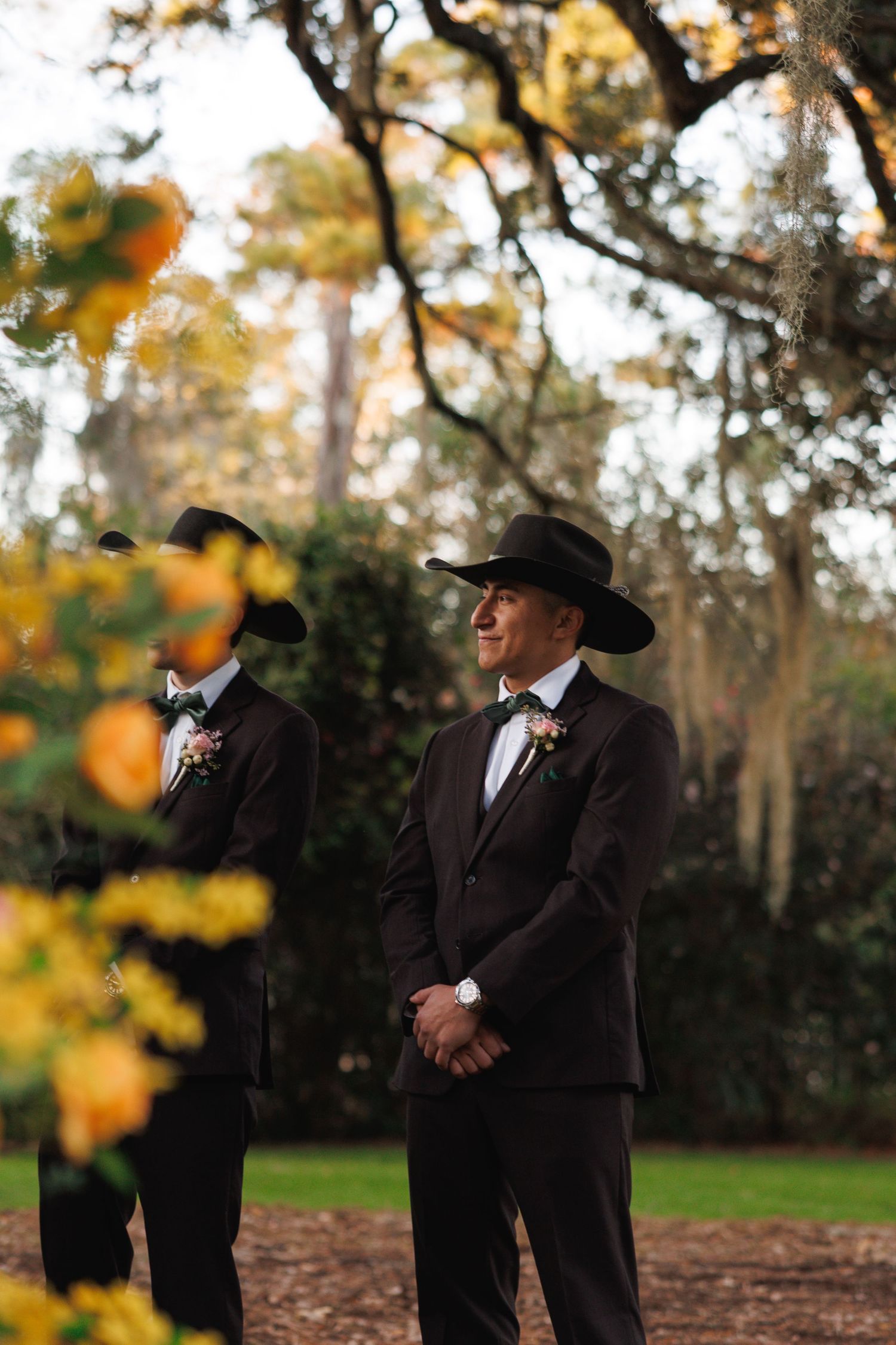 Groomsman in dark suit and black cowboy hat smiling outdoors among autumn foliage at a wedding.