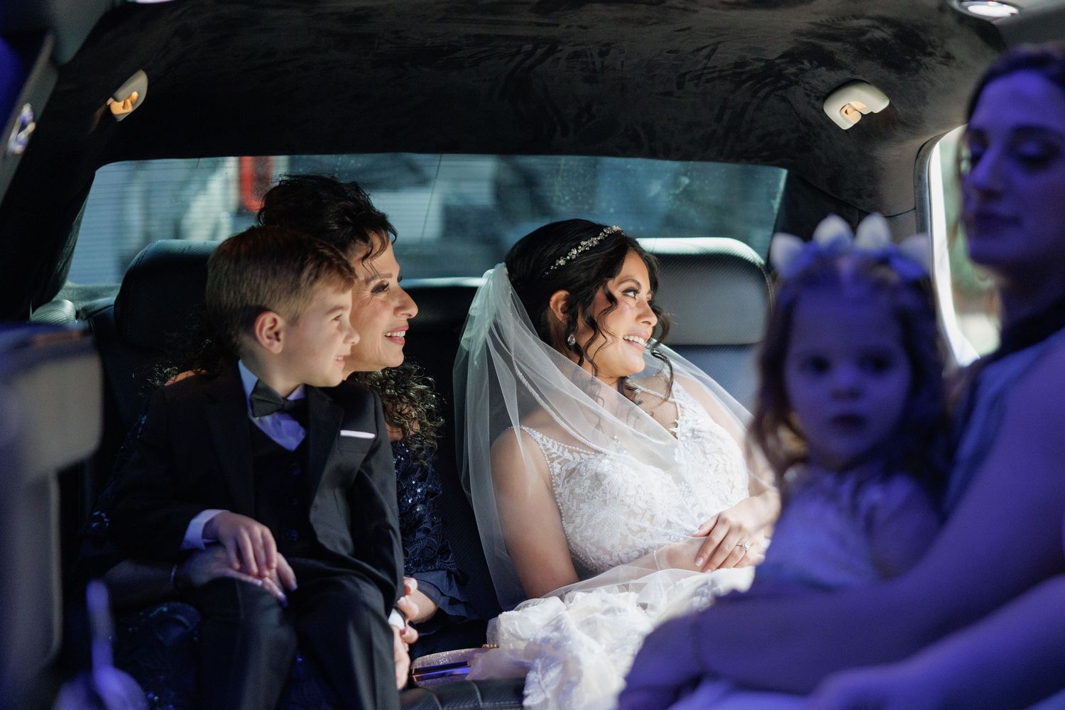 Bride in lace gown and veil sits with wedding party inside a limousine on her wedding day.