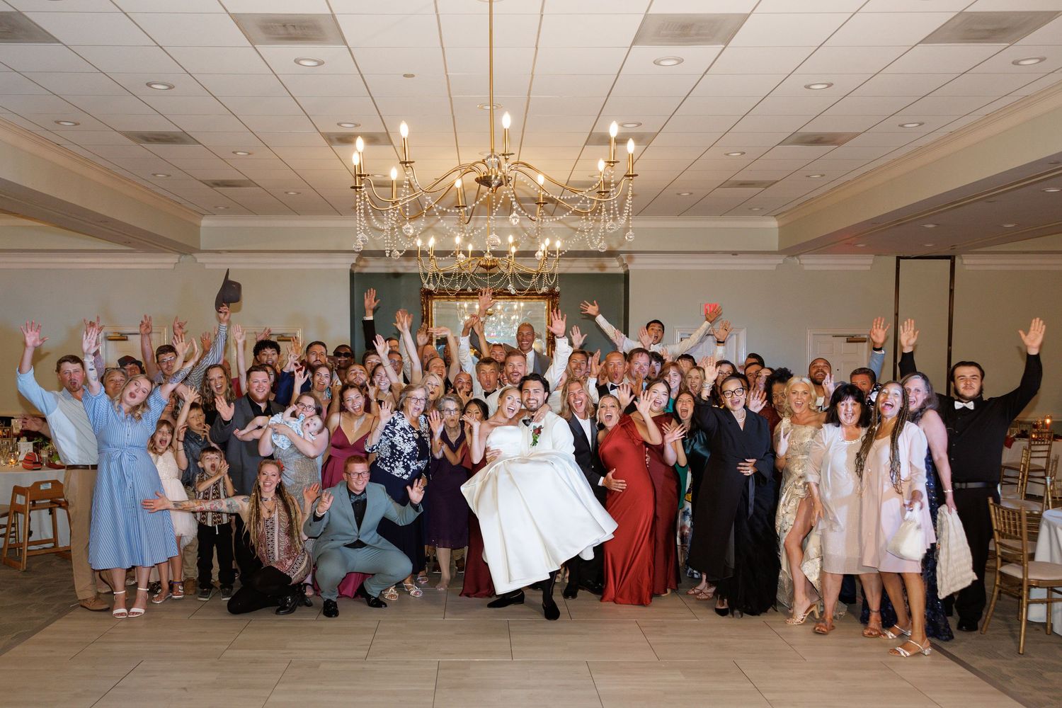Large joyful wedding reception group photo with bride and groom surrounded by celebrating guests in elegant ballroom.