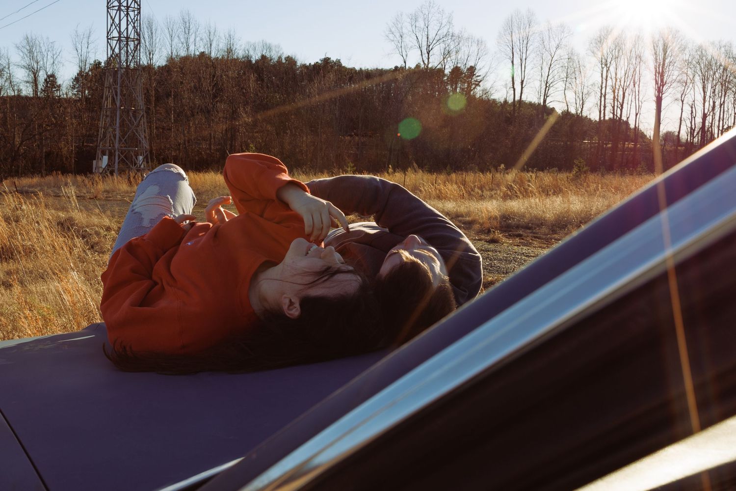 Person in orange hoodie lying on car roof in sunny autumn field with golden grass and trees in background.