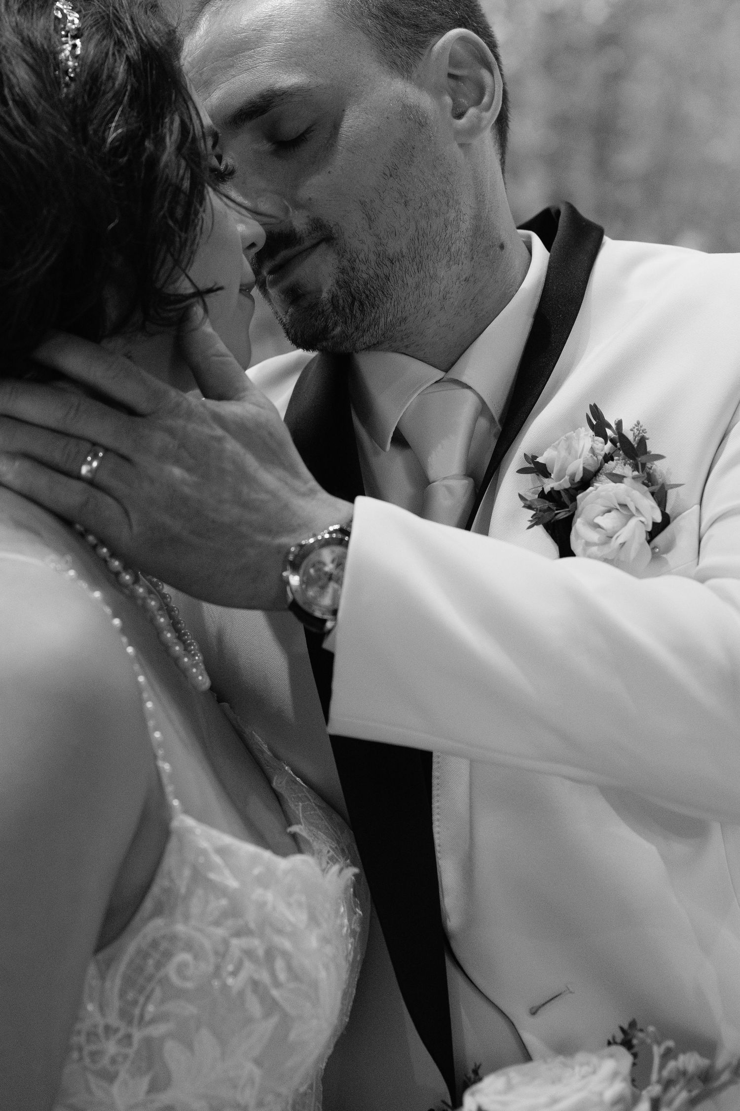 Black and white photo of groom in white tuxedo tenderly kissing bride on wedding day.