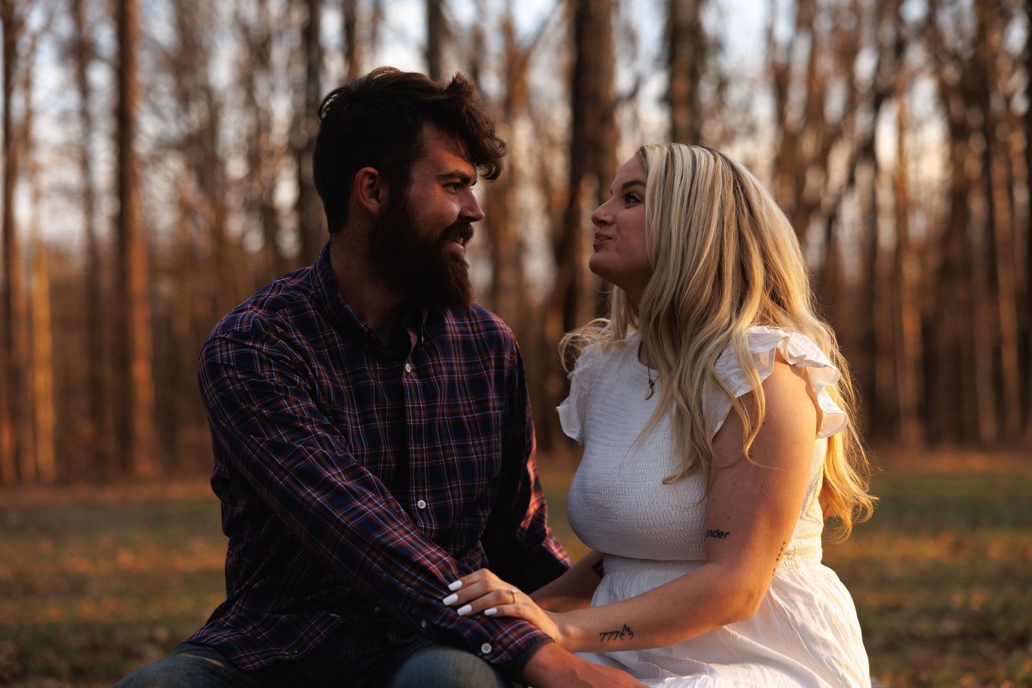 A bearded man in plaid and blonde woman in white dress gaze at each other in an autumn forest.