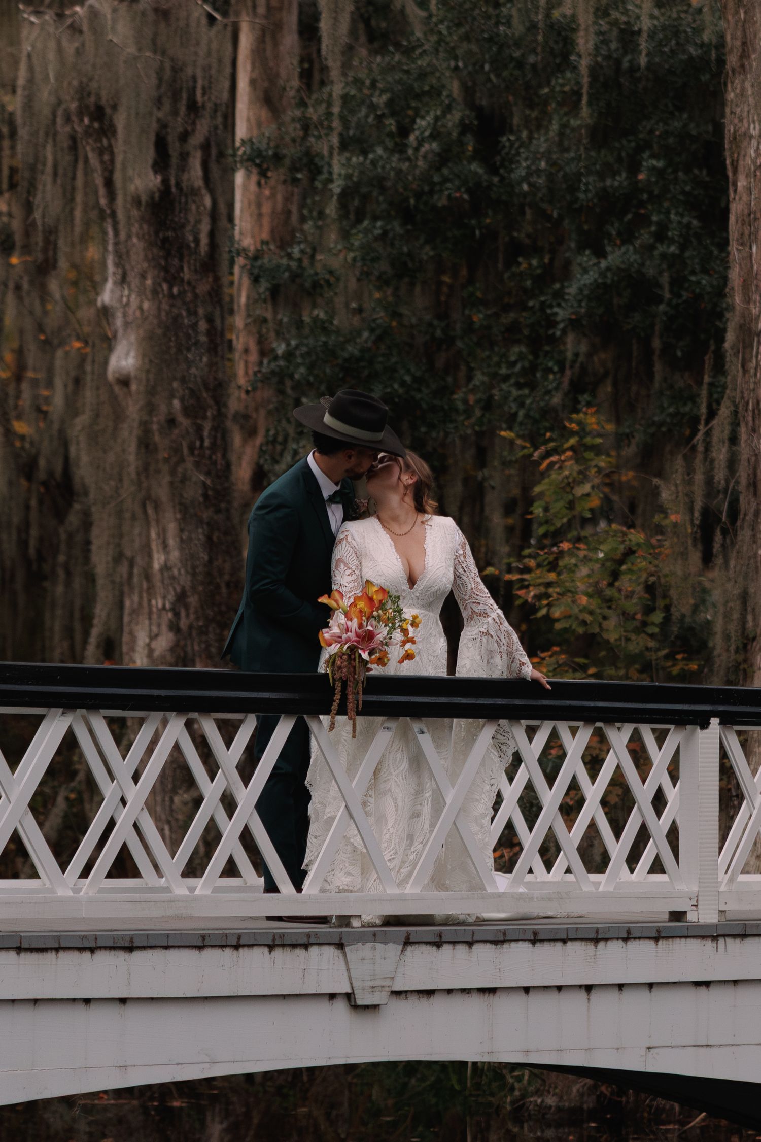 Bride and groom kissing on white ornate bridge surrounded by moss-draped trees holding colorful bouquet.