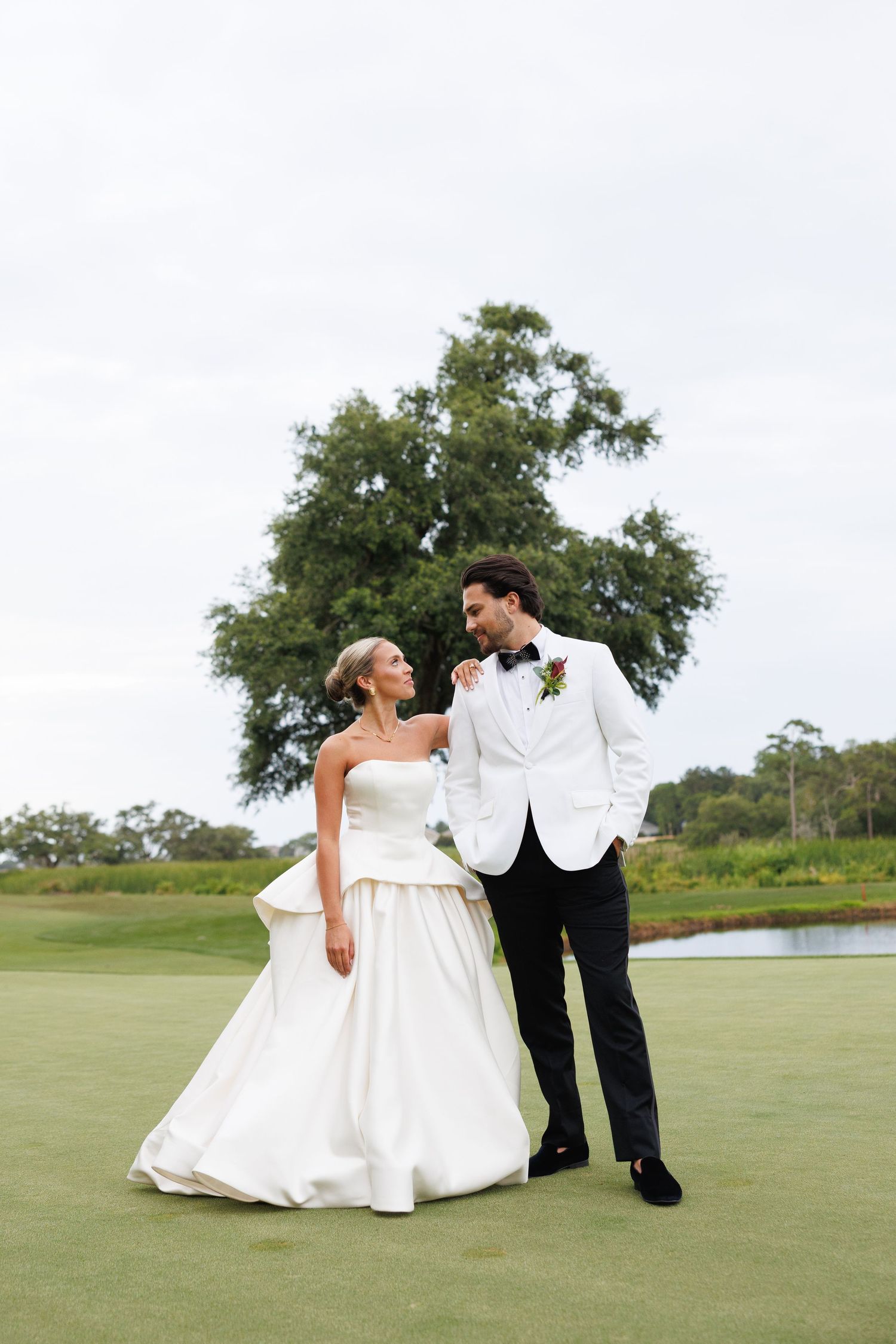 Bride in strapless white ball gown and groom in white tuxedo jacket share a loving glance on a golf course.