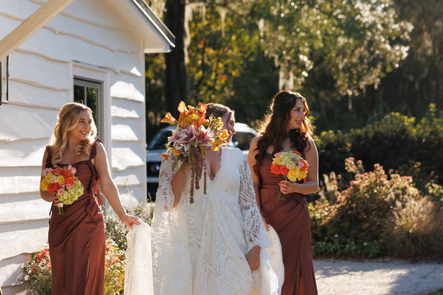Bride in lace gown walks with two bridesmaids in rust dresses holding colorful fall bouquets outside a white chapel.