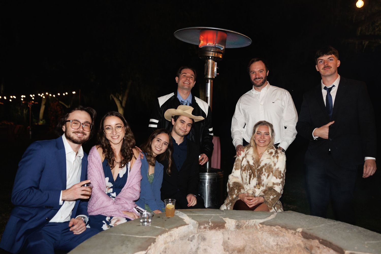 Group of friends posing together outdoors at a night event near a patio heater, dressed in formal attire.
