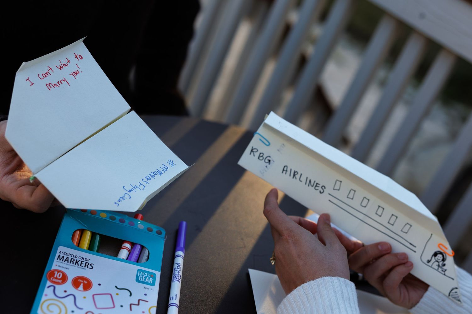 Hands decorating paper airplanes with colorful markers on a table, with a box of Crayola markers nearby.