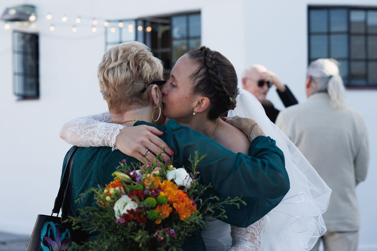 Bride in lace wedding dress embracing a guest while holding colorful bridal bouquet at outdoor venue.