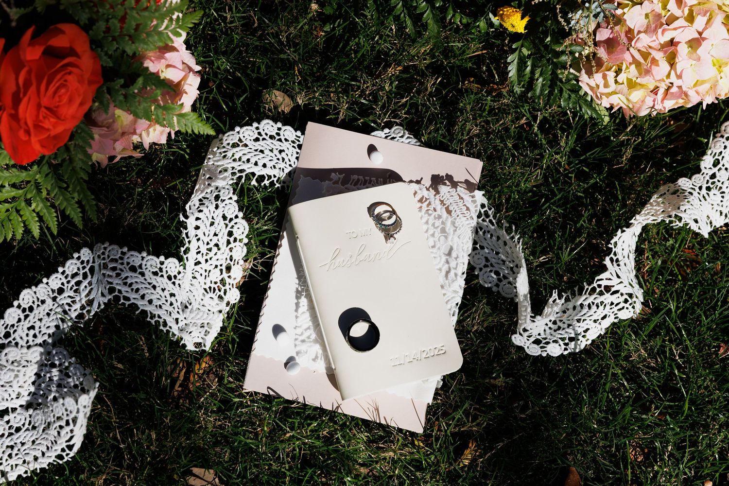 Elegant wedding stationery with lace ribbon on green moss, surrounded by flowers.
