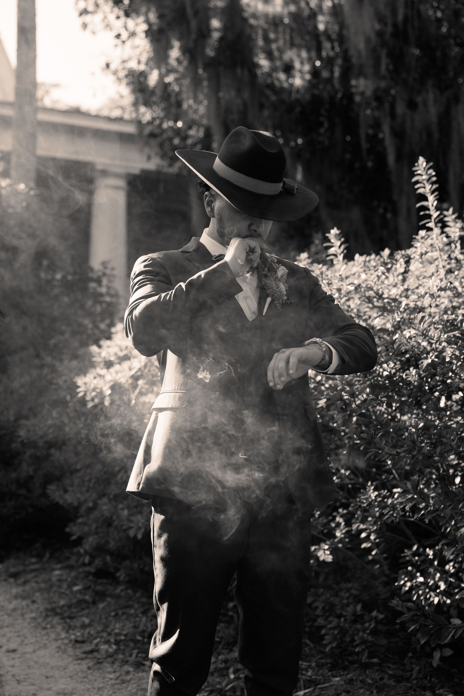 Black and white photo of a groom in a suit and fedora checking his watch amid smoke in a garden.