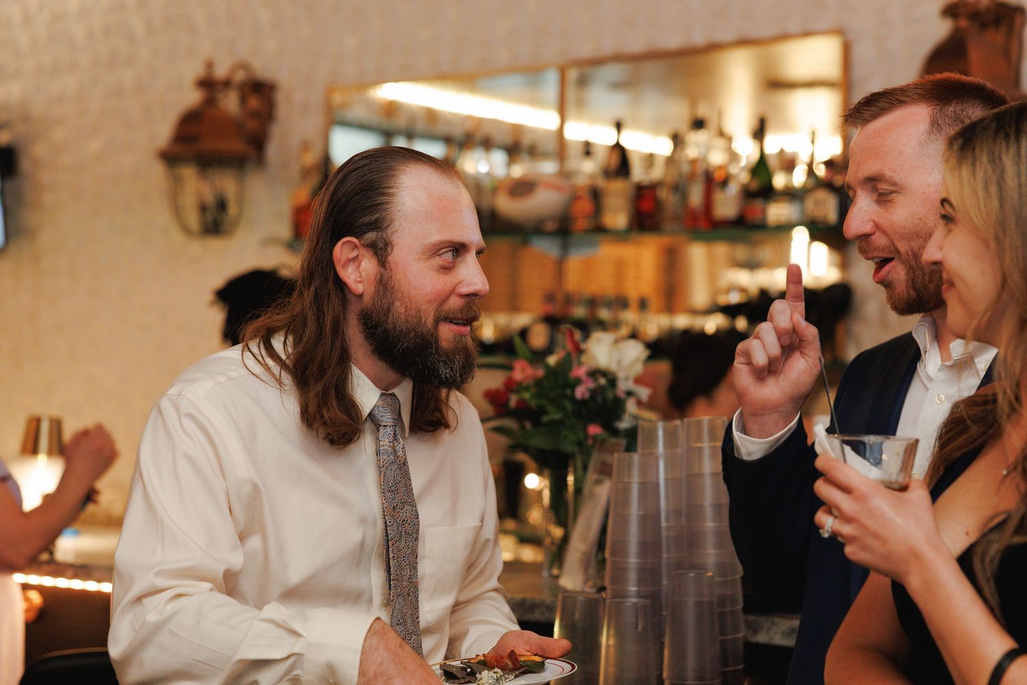 Guests laugh and socialize at wedding reception bar, man with beard and tie enjoys conversation.