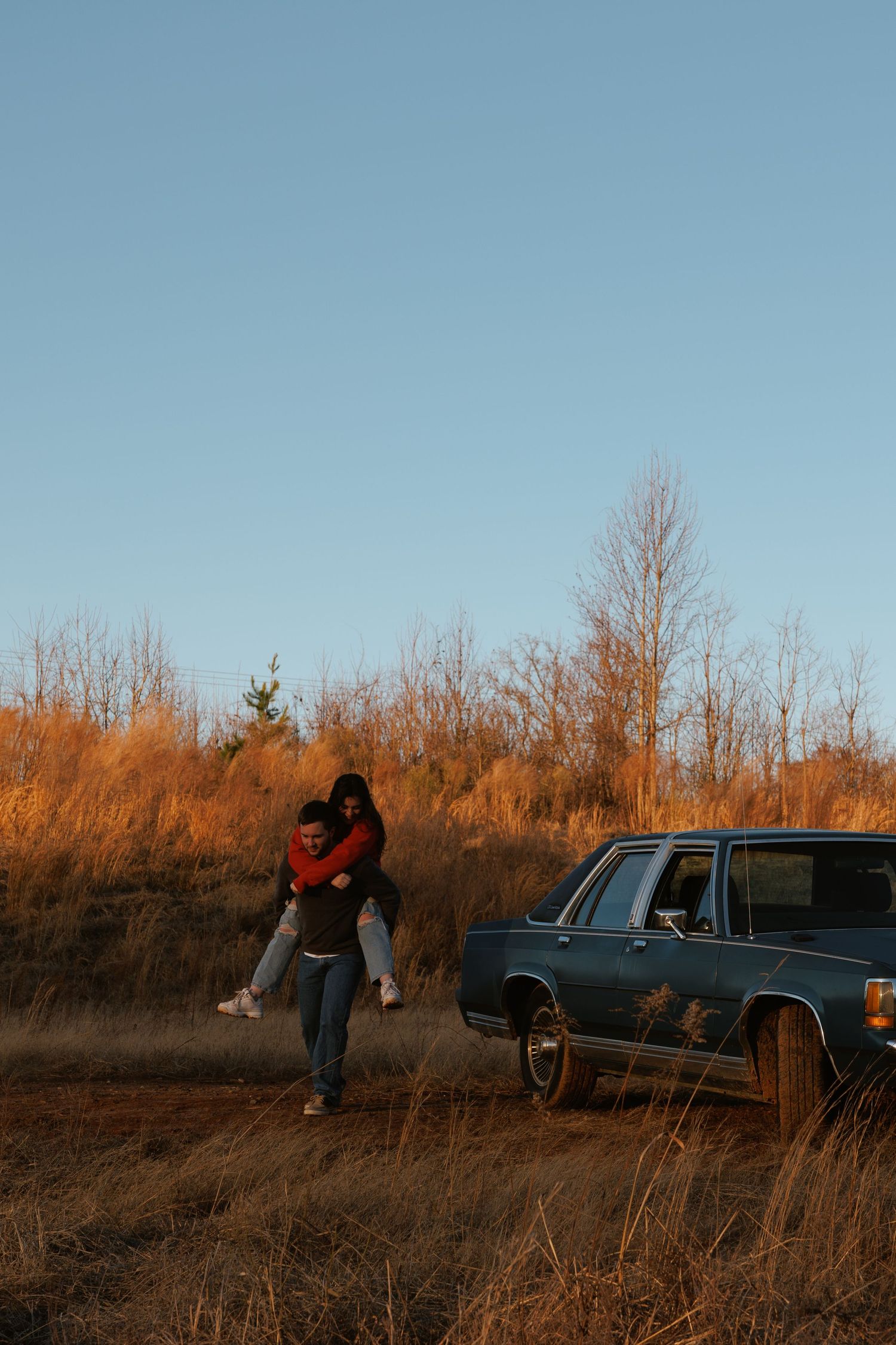 A couple sharing a piggyback ride near a vintage blue car in a golden autumn field.