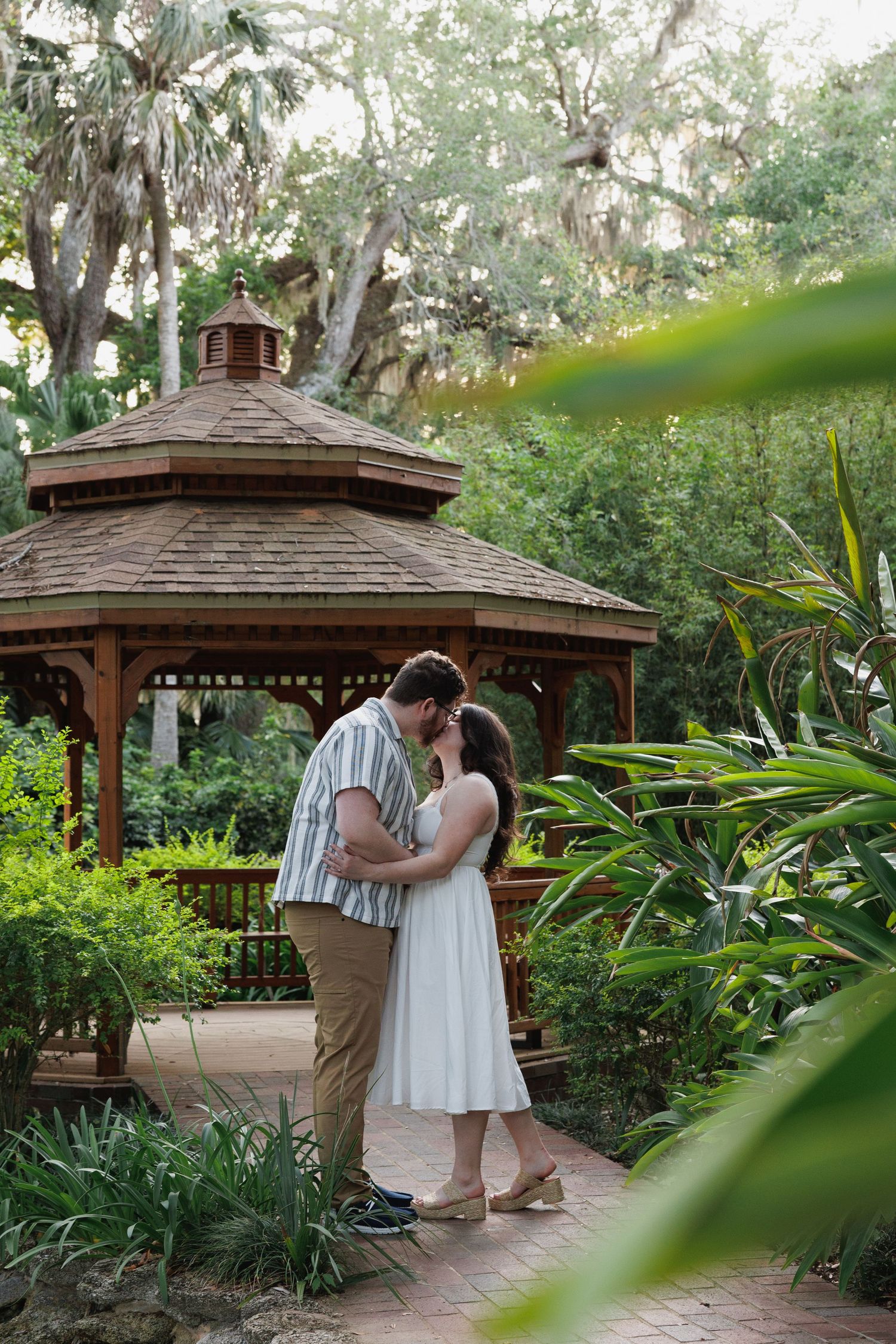Couple sharing a romantic kiss in front of a wooden gazebo surrounded by lush tropical greenery.