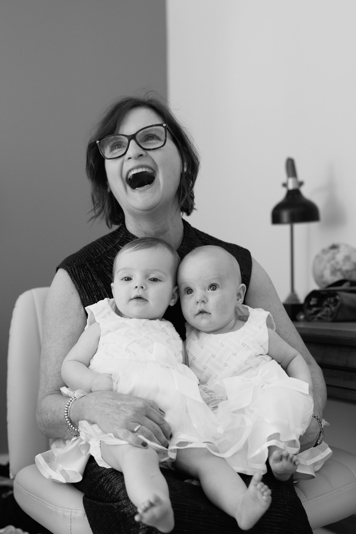 Laughing woman with glasses holds two adorable babies in white dresses in black and white portrait photo.