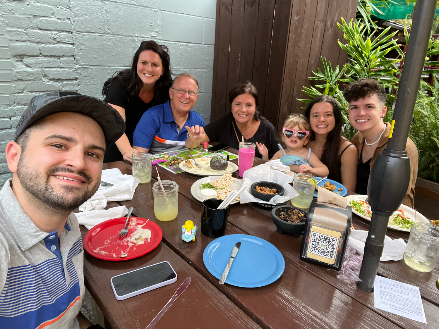 Group of seven friends smiling and posing for a selfie at an outdoor restaurant table with food and drinks.