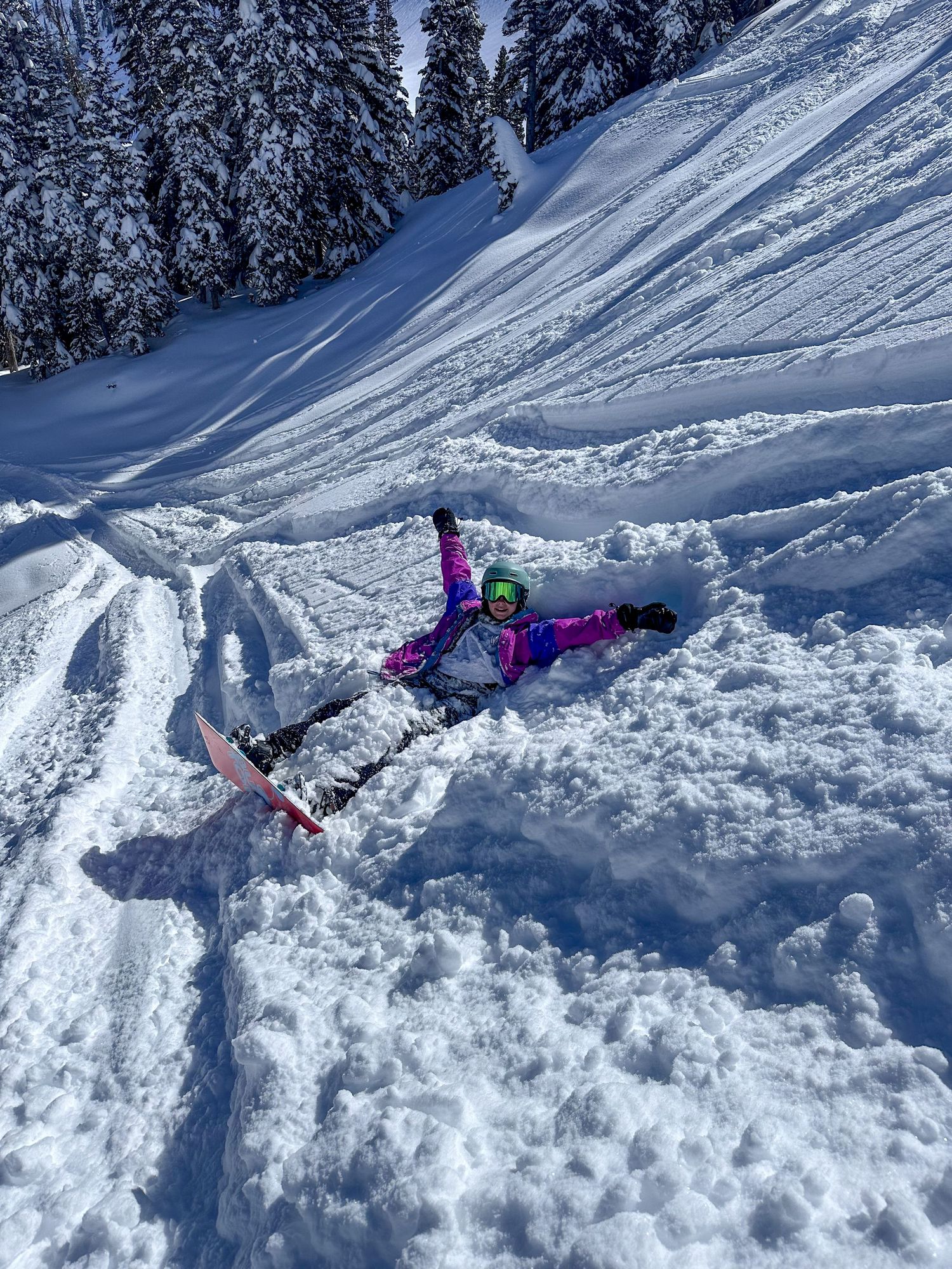 A snowboarder in bright pink gear laughs while lying in deep powder snow on a sunny mountain slope.