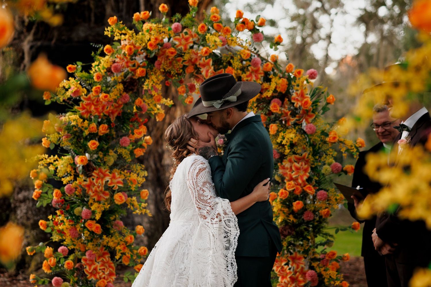 Couple sharing first kiss under vibrant orange and yellow floral arch at outdoor wedding ceremony.