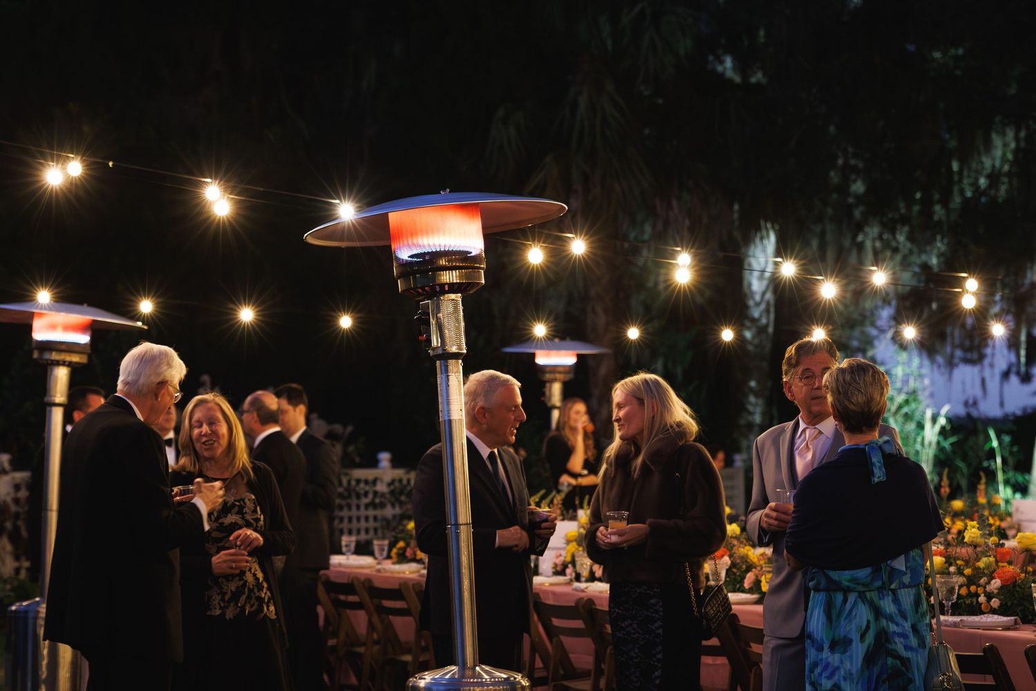Elegant outdoor evening wedding reception with string lights and patio heaters, guests mingling in formal attire.