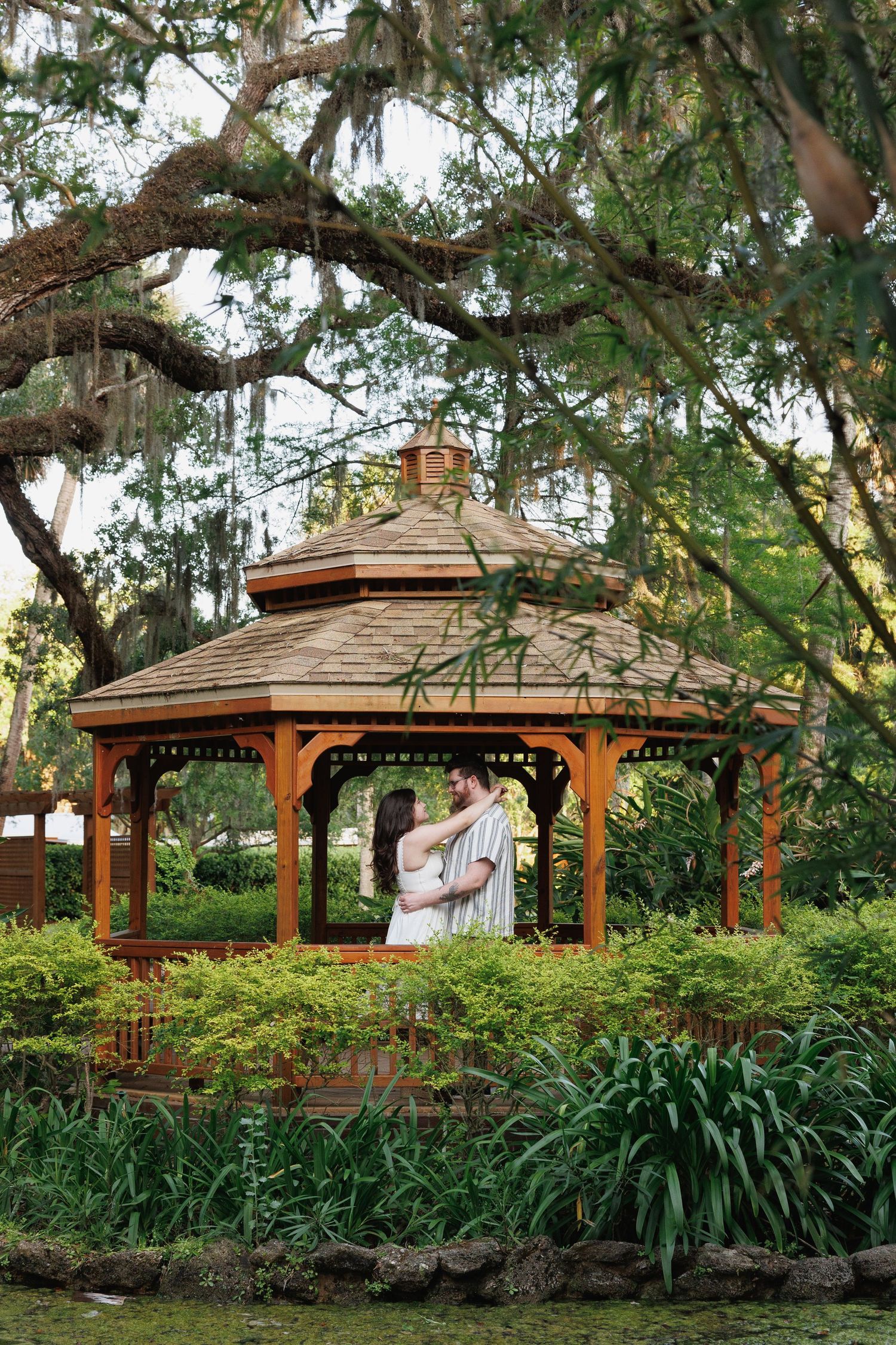 Couple embracing in a wooden gazebo surrounded by lush greenery and Spanish moss-draped oak trees in a garden. engagement session in Jax, fl