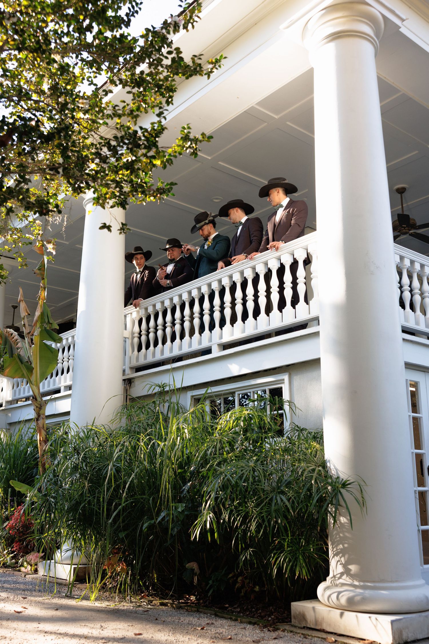 Groomsmen in dark suits and cowboy hats standing on white colonial mansion balcony.