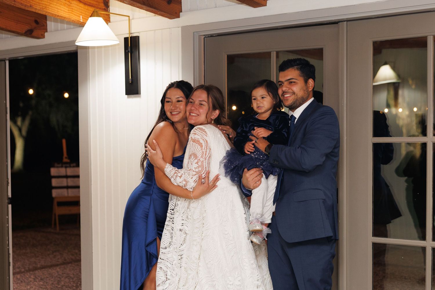Bride in white lace gown embracing family members including a young child and man in navy suit at wedding venue.